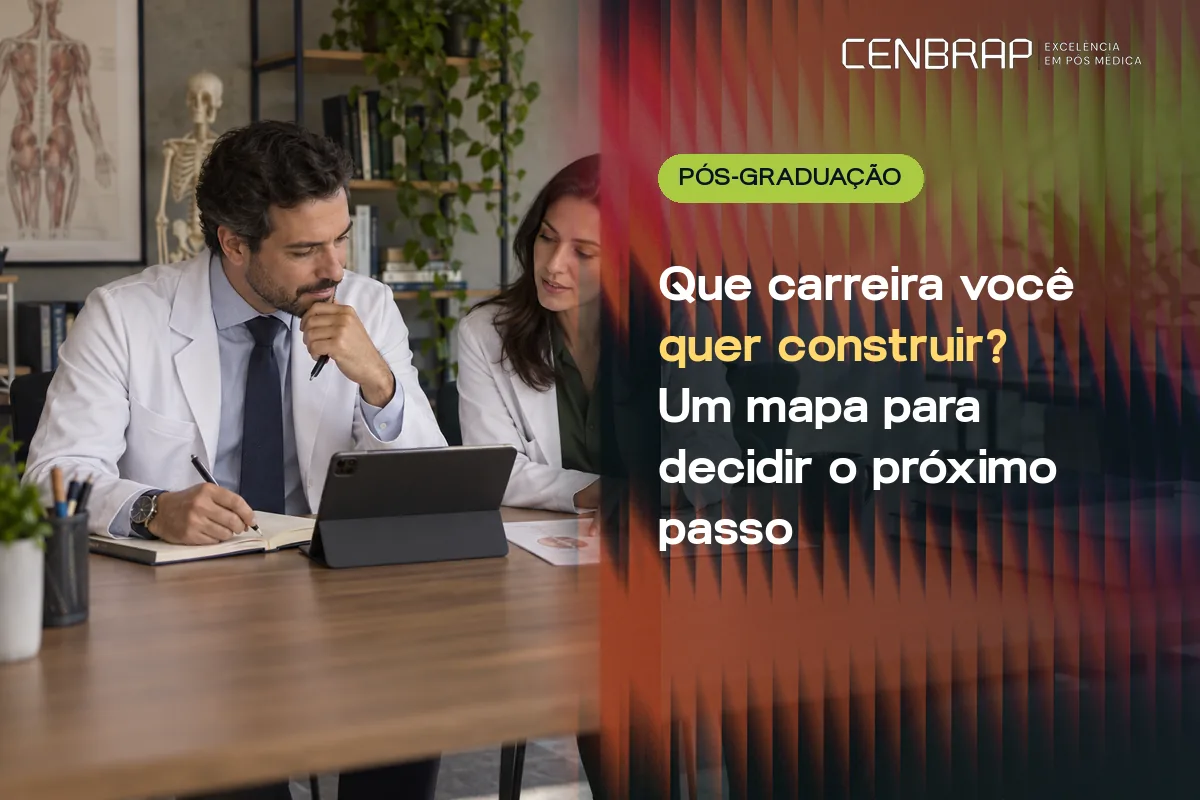 Médico e médica de jaleco sentados lado a lado a uma mesa, conversando sobre anotações e um tablet, com estantes e um modelo anatômico ao fundo.